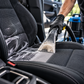 A close-up of a car seat being cleaned with a handheld vacuum attachment. A person wearing a black glove is visible, along with part of a blue and black cleaning machine in the background inside a vehicle.